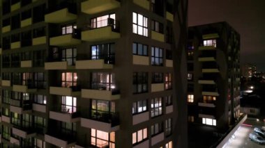 Karanlık bir gökyüzünün altında kentsel manzarayla çevrili aydınlık pencereleri ve balkonları olan modern bir apartmanın gece manzarası..