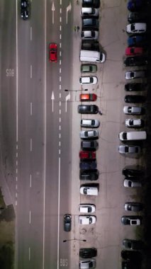 Gece bir şehir sokağının yanındaki park alanının hava görüntüsü, park halindeki arabalar ve trafik ışıklarıyla aydınlatılıyor..