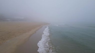 Yumuşak kum ve kıyıya vuran hafif dalgalarla sisli bir sahil. Kıyı boyunca ayak izleri var, sisli atmosfere karışıyorlar. Sakin su ve sessiz tonlar huzurlu ve sakin bir manzara yaratır..