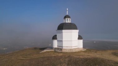 Bulgaristan 'daki küçük beyaz bir kilise, sis ve geniş bir manzarayla çevrili bir tepenin üzerinde duruyor. Kara kubbeleri ve haçı, mavi gökyüzüne ve sisli vadiye karşı dingin, ruhani bir atmosfer yaratır..