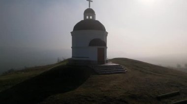 Bulgaristan 'daki küçük beyaz bir kilise, sis ve geniş bir manzarayla çevrili bir tepenin üzerinde duruyor. Kara kubbeleri ve haçı, mavi gökyüzüne ve sisli vadiye karşı dingin, ruhani bir atmosfer yaratır..
