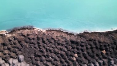 Turkuaz su ile göldeki engebeli arazi arasında çarpıcı bir zıtlık var. Endüstriyel alan, keskin dokuları canlı renklerle harmanlayarak çevresel etkinin katmanlarını ortaya çıkarıyor..