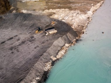 Turkuaz göl kenarında ağır makineleri olan bir çimento fabrikası. Kamyon ve buldozerler, endüstriyel süreçleri vurgulayarak, maddeyi çorak, gri arazide taşıyorlar.