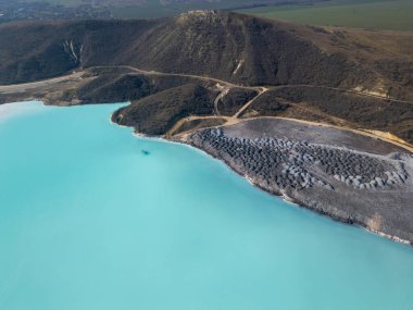 A turquoise tailings pond near a cement factory, surrounded by rugged hills. The vivid water contrasts with the barren land, creating a striking yet industrial scene of environmental impact. clipart