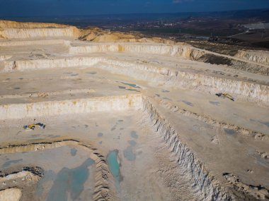 Tozlu arazide çalışan teraslı katmanlı ve ağır makineli bir açık maden ocağı. Sanayi sahasında geniş çaplı kazı süreçleri ve çevresel dönüşüm vurgulanıyor.