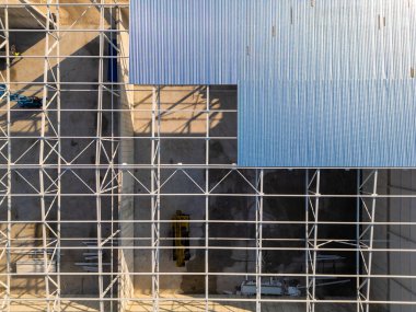 Çelik iskeleti ve metal çatısı olan kısmen inşa edilmiş bir depo. İşçiler ve ağır makineler sanayi ilerlemesi ve yapısal gelişimi gösteren mekanda faaliyet gösteriyorlar.