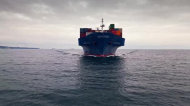 Bulutlu bir gökyüzünün altında durgun sularda seyreden mavi bir konteynır gemisi limana doğru ilerliyor..