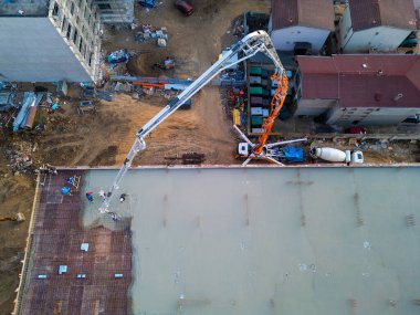 Yüksek binası olan bir inşaat alanının havadan görüntüsü. Bir beton pompa çatıya çimento dökerken, işçiler ve iskeleler ilerlemeyi gösteriyor. Etrafı modern insanlarla çevrili