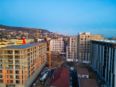 Yüksek binası olan bir inşaat alanının havadan görüntüsü. Bir beton pompa çatıya çimento dökerken, işçiler ve iskeleler ilerlemeyi gösteriyor. Etrafı modern insanlarla çevrili