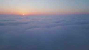 Geniş bir bulut denizinin üzerinde gün doğumu. Altın ışık, sisli gökyüzünü nazikçe aydınlatır. Yumuşak renkler ve yumuşak hareketlerle rüya gibi, sakin bir atmosfer yaratır. Hava aracı görünümü.