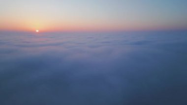 Geniş bir bulut denizinin üzerinde gün doğumu. Altın ışık, sisli gökyüzünü nazikçe aydınlatır. Yumuşak renkler ve yumuşak hareketlerle rüya gibi, sakin bir atmosfer yaratır. Hava aracı görünümü.