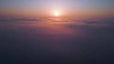 Geniş bir bulut denizinin üzerinde gün doğumu. Altın ışık, sisli gökyüzünü nazikçe aydınlatır. Yumuşak renkler ve yumuşak hareketlerle rüya gibi, sakin bir atmosfer yaratır. Hava aracı görünümü.