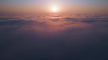 Geniş bir bulut denizinin üzerinde gün doğumu. Altın ışık, sisli gökyüzünü nazikçe aydınlatır. Yumuşak renkler ve yumuşak hareketlerle rüya gibi, sakin bir atmosfer yaratır. Hava aracı görünümü.