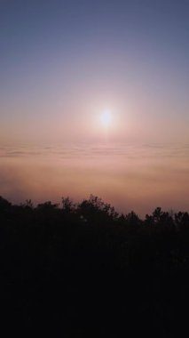 Puslu bir dağ manzarası üzerinde büyüleyici bir gün doğumu. Altın ışık, yuvarlanan bulutları aydınlatıyor. Sakin atmosfer, orman siluetlerini yumuşak sabah renkleriyle harmanlıyor ve nefes kesici bir hava yaratıyor.