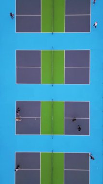 Oyuncuların bir maçta yer aldığı çoklu picleball sahalarının yukarıdan aşağı görüntüsü. Canlı mavi ve yeşil yüzeyler çarpıcı bir geometrik desen yaratarak dinamik hareketi ve enerjiyi yakalıyor.