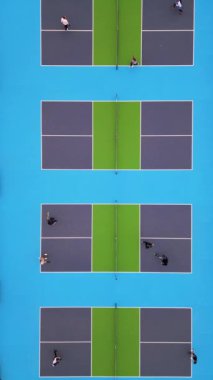 Oyuncuların bir maçta yer aldığı çoklu picleball sahalarının yukarıdan aşağı görüntüsü. Canlı mavi ve yeşil yüzeyler çarpıcı bir geometrik desen yaratarak dinamik hareketi ve enerjiyi yakalıyor.