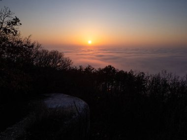 Puslu bir dağ manzarası üzerinde büyüleyici bir gün doğumu. Altın ışık, yuvarlanan bulutları aydınlatıyor. Sakin atmosfer, orman siluetlerini yumuşak sabah renkleriyle harmanlıyor ve nefes kesici bir hava yaratıyor.
