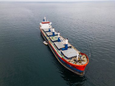 Açık denizde kızıl bir pruvası olan büyük bir kargo gemisi, bulutlu bir gökyüzünün altında bir römorkör eşliğinde, deniz taşımacılığı sahnesi..