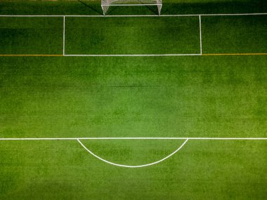 Yeşil suni çim, beyaz kale direği ve parlak ışıklandırma altında görünen alan işaretleri olan bir futbol sahasının yukarıdan aşağıya havadan görünüşü..