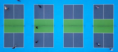 Oyuncuların bir maçta yer aldığı çoklu picleball sahalarının yukarıdan aşağı görüntüsü. Canlı mavi ve yeşil yüzeyler çarpıcı bir geometrik desen yaratarak dinamik hareketi ve enerjiyi yakalıyor.
