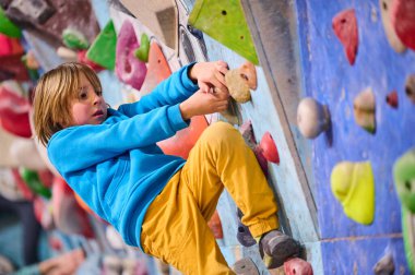 Mavi kapüşonlu bir çocuk, kapalı bir kaya duvarına tırmanıyor, odak ve kararlılıkla renkli tutumlar sergiliyor. Bir sonraki durağa uzanıyor, azim, güç ve macera sergiliyor..