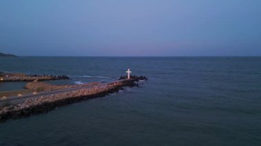 Alacakaranlıkta aydınlanmış deniz feneri kulesi okyanusa bakıyor, uzak bir iskelede ışıldayan bir haç var. Yumuşak pastel gökyüzü ve ışığı yansıtan barışçıl dalgalarla sakin bir deniz manzarası..