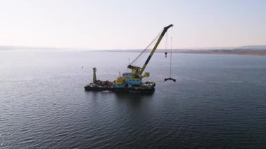 Büyük sarı bir vinci olan yüzen bir vinç sakin sularda çalışır ve kancasıyla bir nesneyi kaldırır. Endüstriyel gemi uzak bir kıyı şeridiyle çevrili..