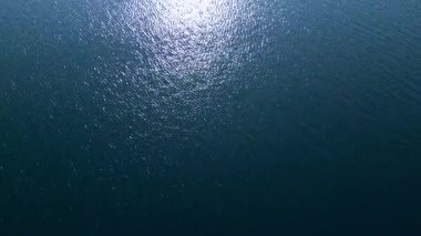 Durgun bir su yüzeyinin hava görüntüsü. Güneş ışığı yansımaları ışıltılı bir etki yaratıyor. Yumuşak dalgalar, derin mavi alana dokular ekler. Huzuru ve doğanın güzelliğini çağrıştırır..