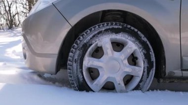 Bir araba, tekerleği derin karda dönerken hareket etmeye çabalıyor. Araç kısmen sıkışmış durumda. Lastik buz ve karla kaplı. Bu da kış yolculuğunun ve yol koşullarının zorluklarını gösteriyor..