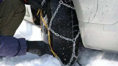 Eldiven giyen biri buzlu yollarda daha iyi yol tutuşu için araba lastiğine kar zinciri takar. Yakın plan çekimde metal halkalar ve sert kış lastikleri ön plana çıkıyor..