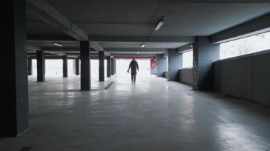 Yalnız bir adam loş ışıklı, beton sütunları ve açık pencereleri olan boş bir otoparkta yürür ve gölgeler bırakır. Endüstriyel alanda gizemli bir atmosfer var..