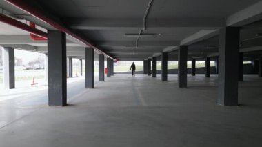 Yalnız bir adam loş ışıklı, beton sütunları ve açık pencereleri olan boş bir otoparkta yürür ve gölgeler bırakır. Endüstriyel alanda gizemli bir atmosfer var..
