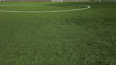 Boş bir futbol sahasının düşük irtifa drone görüntüsü suni yeşil çimenler, beyaz sınır çizgileri ve gol ağı. Spor sahası iyi korunmuş, temiz ve yapılandırılmış bir kompozisyon oluşturuyor..