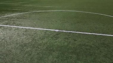 Boş bir futbol sahasının düşük irtifa drone görüntüsü suni yeşil çimenler, beyaz sınır çizgileri ve gol ağı. Spor sahası iyi korunmuş, temiz ve yapılandırılmış bir kompozisyon oluşturuyor..