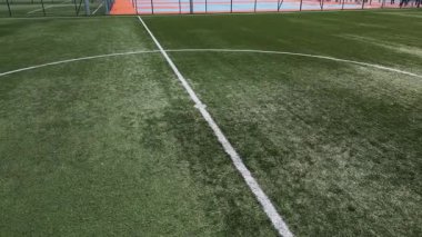 Boş bir futbol sahasının düşük irtifa drone görüntüsü suni yeşil çimenler, beyaz sınır çizgileri ve gol ağı. Spor sahası iyi korunmuş, temiz ve yapılandırılmış bir kompozisyon oluşturuyor..