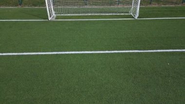 Boş bir futbol sahasının düşük irtifa drone görüntüsü suni yeşil çimenler, beyaz sınır çizgileri ve gol ağı. Spor sahası iyi korunmuş, temiz ve yapılandırılmış bir kompozisyon oluşturuyor..