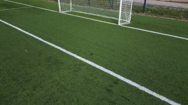 Boş bir futbol sahasının düşük irtifa drone görüntüsü suni yeşil çimenler, beyaz sınır çizgileri ve gol ağı. Spor sahası iyi korunmuş, temiz ve yapılandırılmış bir kompozisyon oluşturuyor..