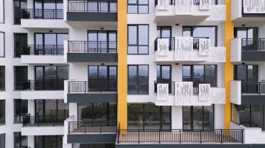 Camdan balkonları ve minimalist tasarımı olan, insansız hava aracı tarafından ele geçirilmiş modern bir yerleşim yeri. Arka planda, vinçleri ve tamamlanmamış bir yapısı olan bir inşaat alanı görülüyor..