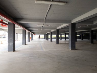 Yalnız bir adam loş ışıklı, beton sütunları ve açık pencereleri olan boş bir otoparkta yürür ve gölgeler bırakır. Endüstriyel alanda gizemli bir atmosfer var..