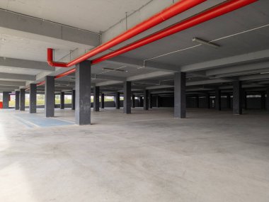 Beton sütunları ve açık kenarları olan boş bir yeraltı otoparkı. Endüstriyel alan doğal ışıkla aydınlanıyor, modern ama terk edilmiş bir atmosfer yaratıyor..
