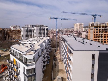 İnşaat alanı, tadilatı devam eden bir bina ve iki büyük vinç. Arka planda yerleşim binaları, yollar ve yeşillik olan şehir manzarası, şehir büyümesini ve