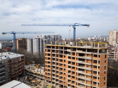 İnşaat alanı, tadilatı devam eden bir bina ve iki büyük vinç. Arka planda yerleşim binaları, yollar ve yeşillik olan şehir manzarası, şehir büyümesini ve