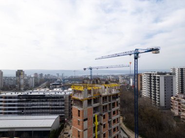 İnşaat alanı, tadilatı devam eden bir bina ve iki büyük vinç. Arka planda yerleşim binaları, yollar ve yeşillik olan şehir manzarası, şehir büyümesini ve