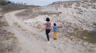 Bir anne ve çocuğu kayalık bir açık araziyi keşfediyorlar. Arazi engebelidir. Arka planda seyrek bitki örtüsü ve uzak tepeler vardır..