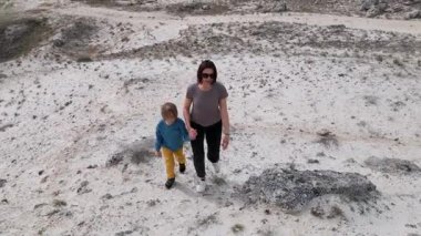 Bir anne ve çocuğu kayalık bir açık araziyi keşfediyorlar. Arazi engebelidir. Arka planda seyrek bitki örtüsü ve uzak tepeler vardır..