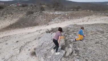 Bir anne ve çocuğu kayalık bir açık araziyi keşfediyorlar. Arazi engebelidir. Arka planda seyrek bitki örtüsü ve uzak tepeler vardır..