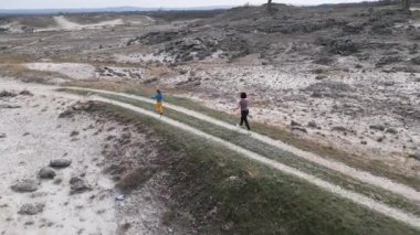 Bir anne ve çocuğu kayalık bir açık araziyi keşfediyorlar. Arazi engebelidir. Arka planda seyrek bitki örtüsü ve uzak tepeler vardır..