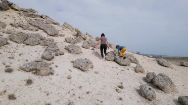 Bir anne ve çocuğu kayalık bir açık araziyi keşfediyorlar. Arazi engebelidir. Arka planda seyrek bitki örtüsü ve uzak tepeler vardır..