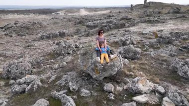 Bir anne ve çocuk engebeli bir doğal ortamda rahatlamak için büyük bir kayanın üzerinde otururlar. Huzurlu ortamın tadını çıkarıyorlar. Dağınık taşlar ve kuru araziler arasında sakin bir anı paylaşıyorlar..