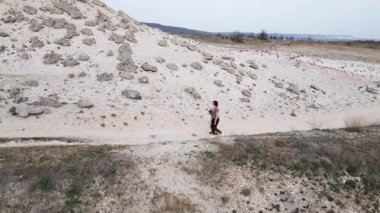 Bir anne ve çocuğu kayalık bir açık araziyi keşfediyorlar. Arazi engebelidir. Arka planda seyrek bitki örtüsü ve uzak tepeler vardır..